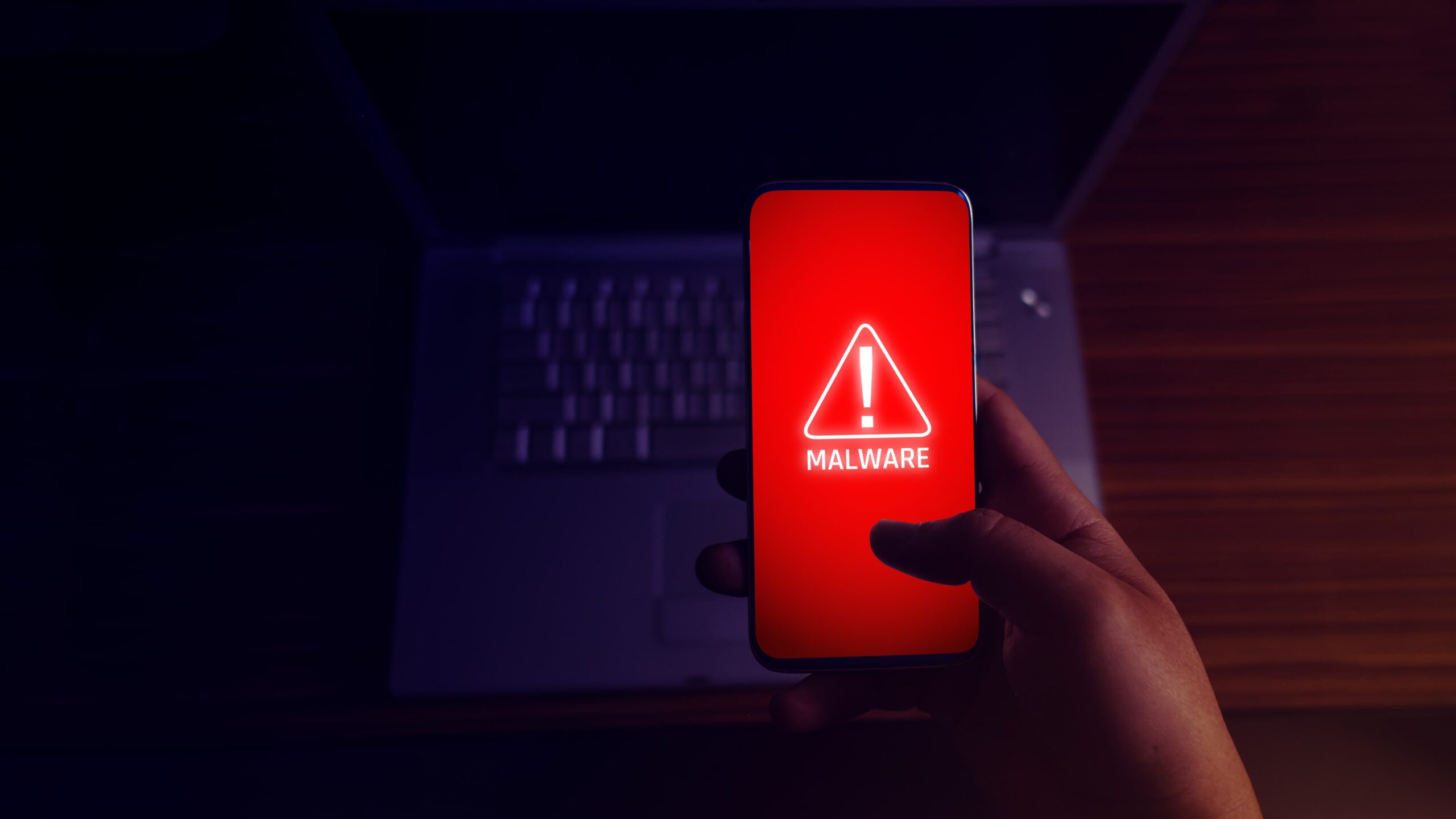 Hand holding a smartphone with a red malware warning screen, indicating a cybersecurity threat, with a laptop in the background.