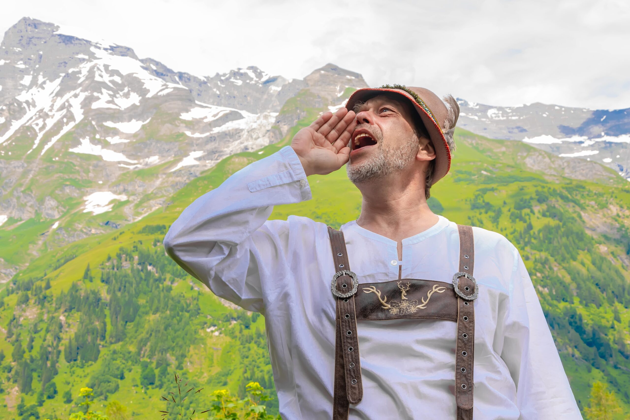 Man in traditional alpine attire yodeling in a mountain landscape with green hills and snow-capped peaks in the background.