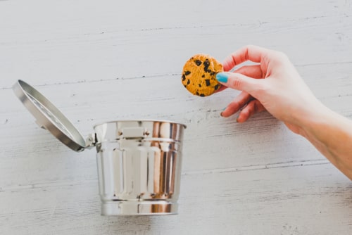 A hand holding a chocolate chip cookie over an open metal trash can on a white wooden surface.