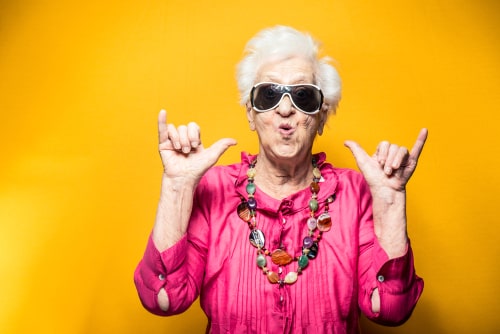 Elderly woman in sunglasses and a bright pink shirt making a fun gesture against a yellow background