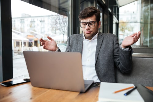 Confused businessman shrugging while looking at laptop screen in a café