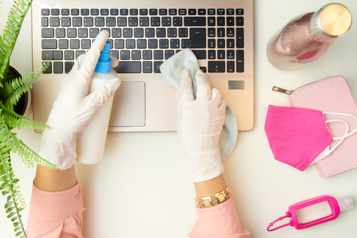 Person wearing gloves cleaning laptop keyboard with disinfectant, surrounded by spring items like face mask and sanitizer