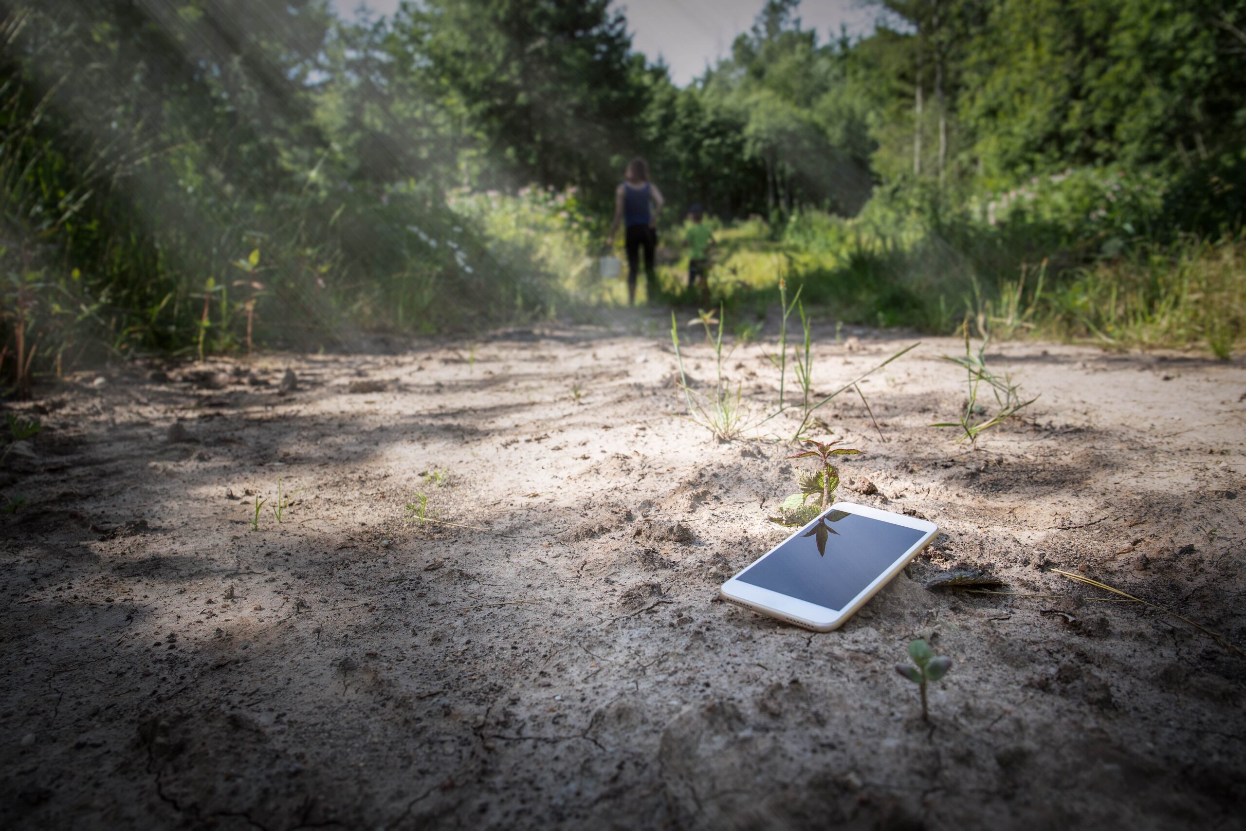 Smartphone lying on dirt path in forest with person walking away in background, representing lost or forgotten phone