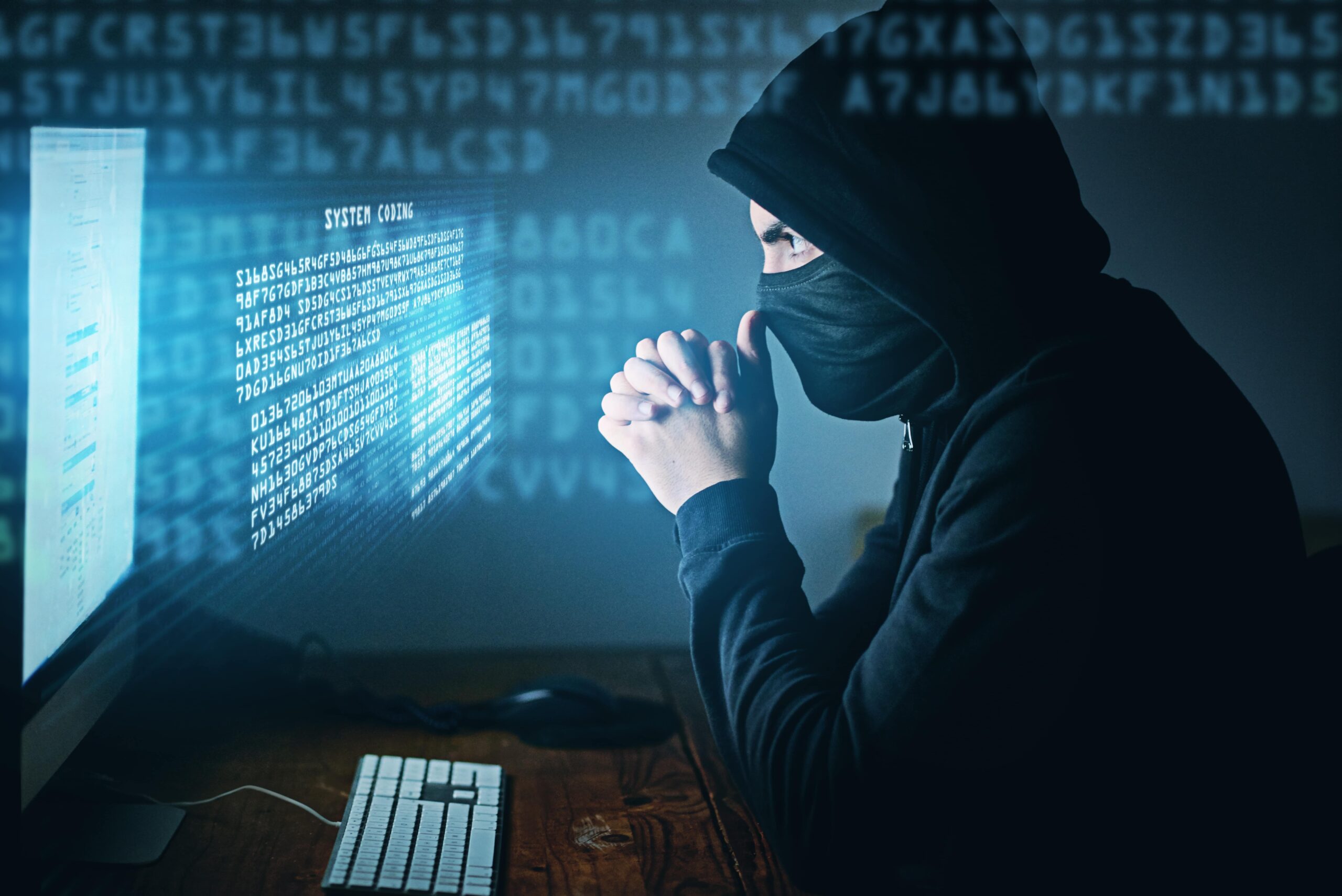 Masked hacker in hoodie working at computer with system coding interface, symbolizing cybercrime or tech scams