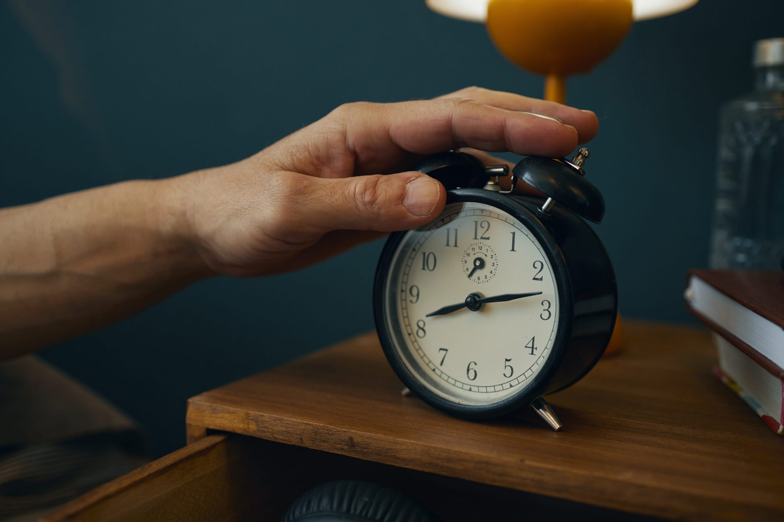 Hand reaching to stop ringing alarm clock on wooden bedside table