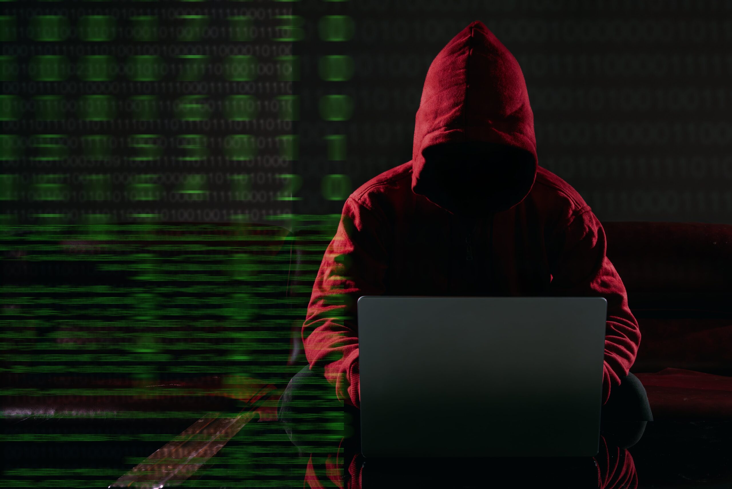 Anonymous hacker in red hoodie using laptop with binary code overlay