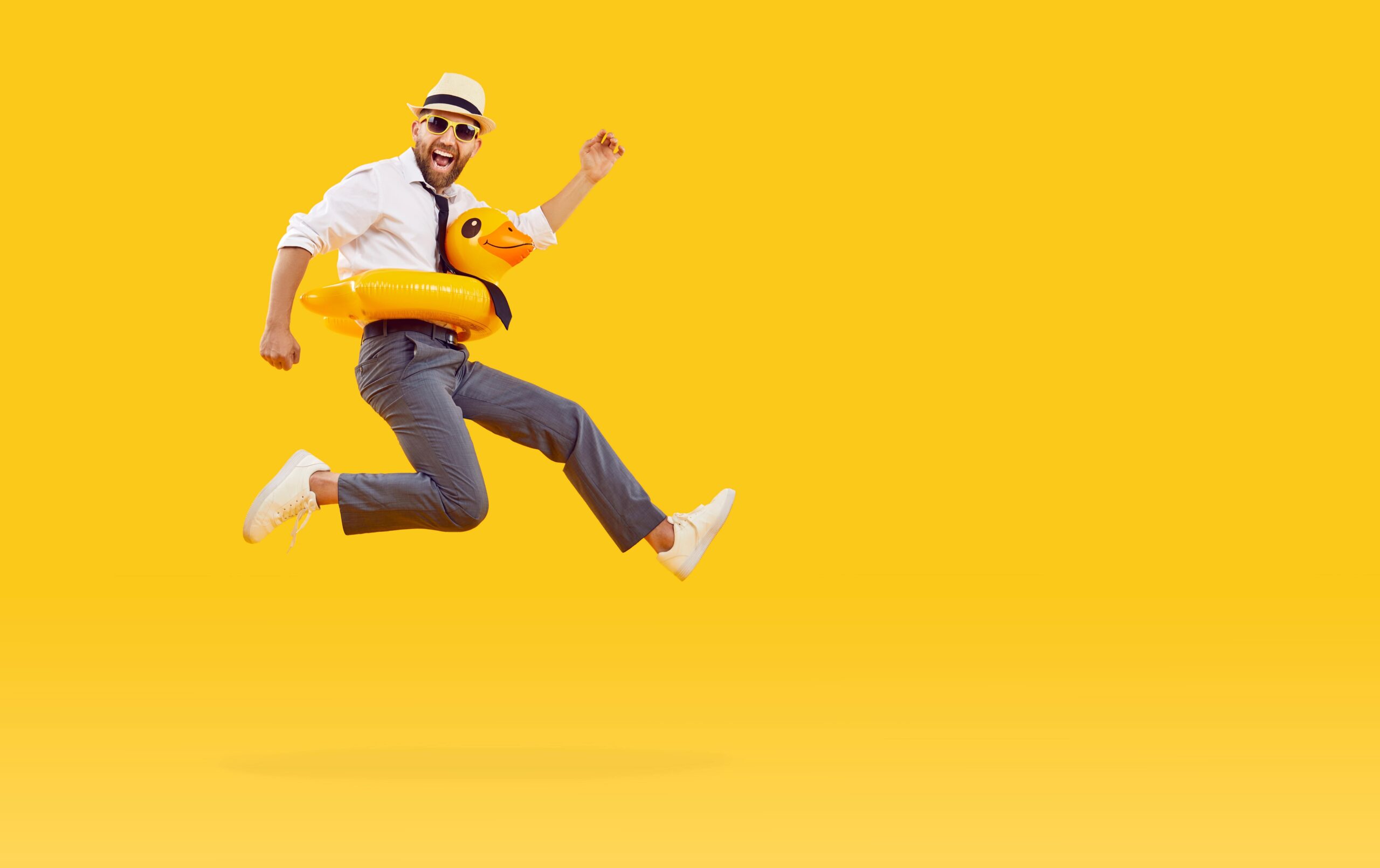 Man in suit, sunglasses, and hat happily jumping with a duck pool floaty on bright yellow background