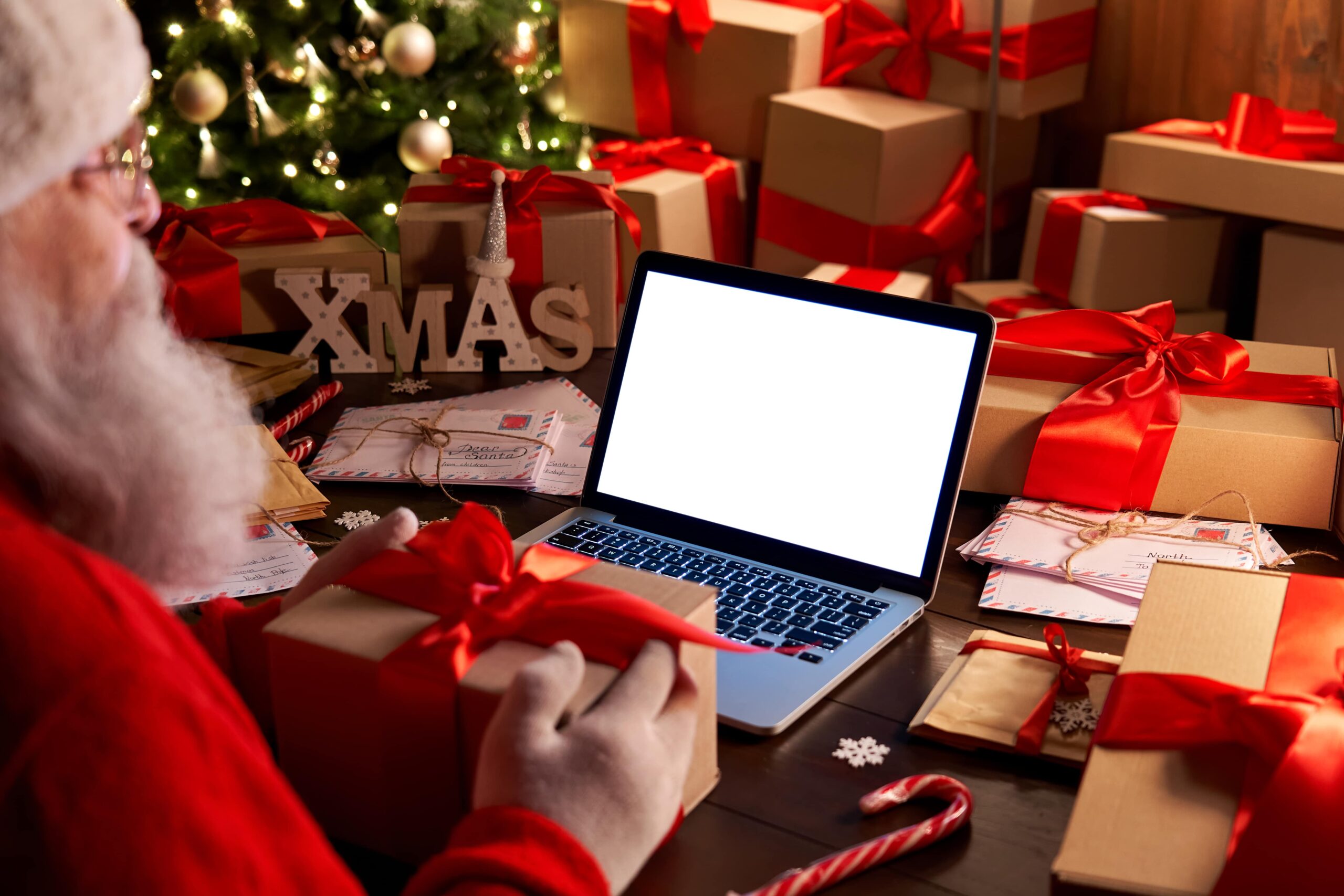 Santa Claus is holding a present in front of a laptop that is displaying a white screen.