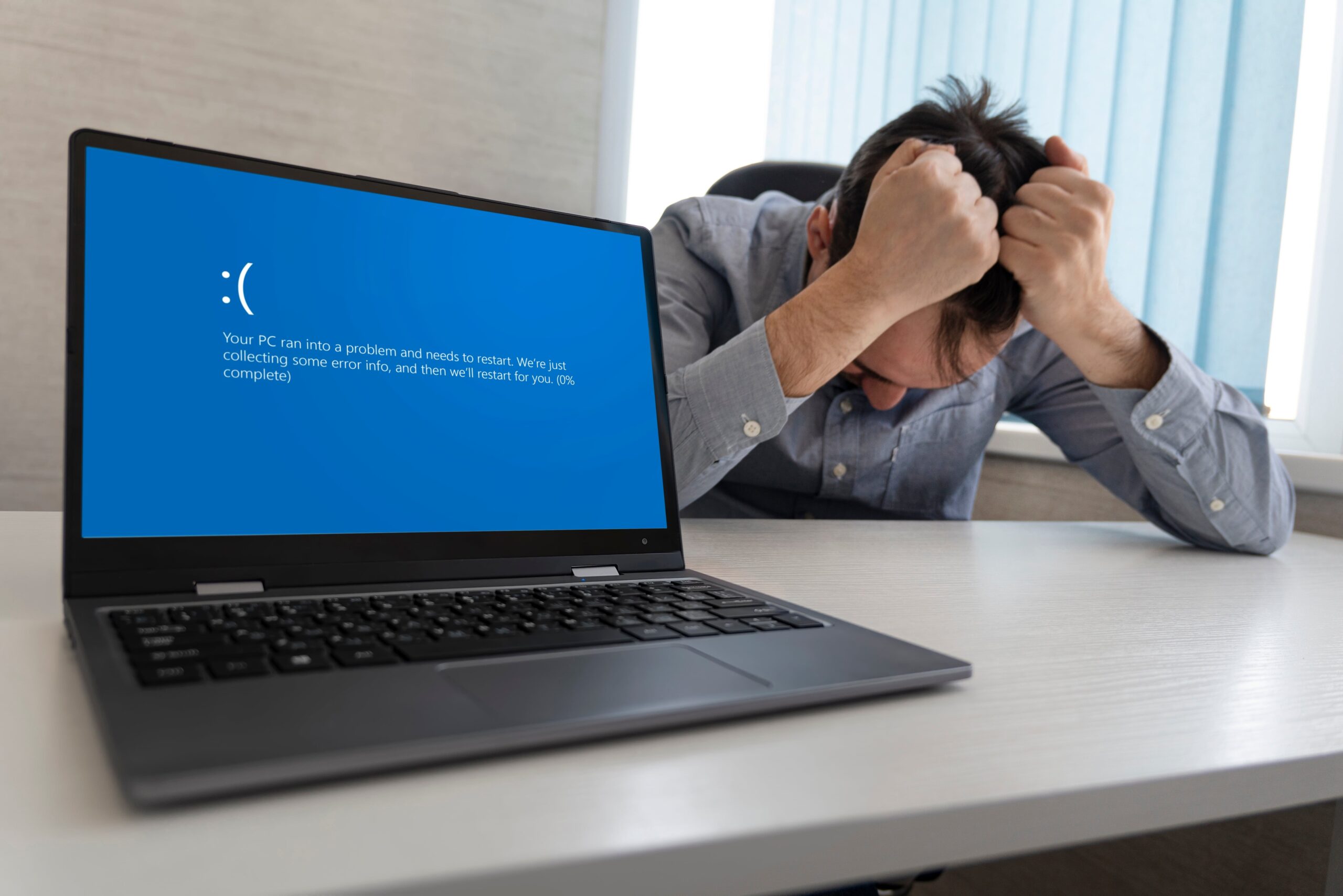 man holding his head behind a Windows computer showing a message saying, “Your PC ran into a problem that it couldn’t handle, and now it needs to restart.”