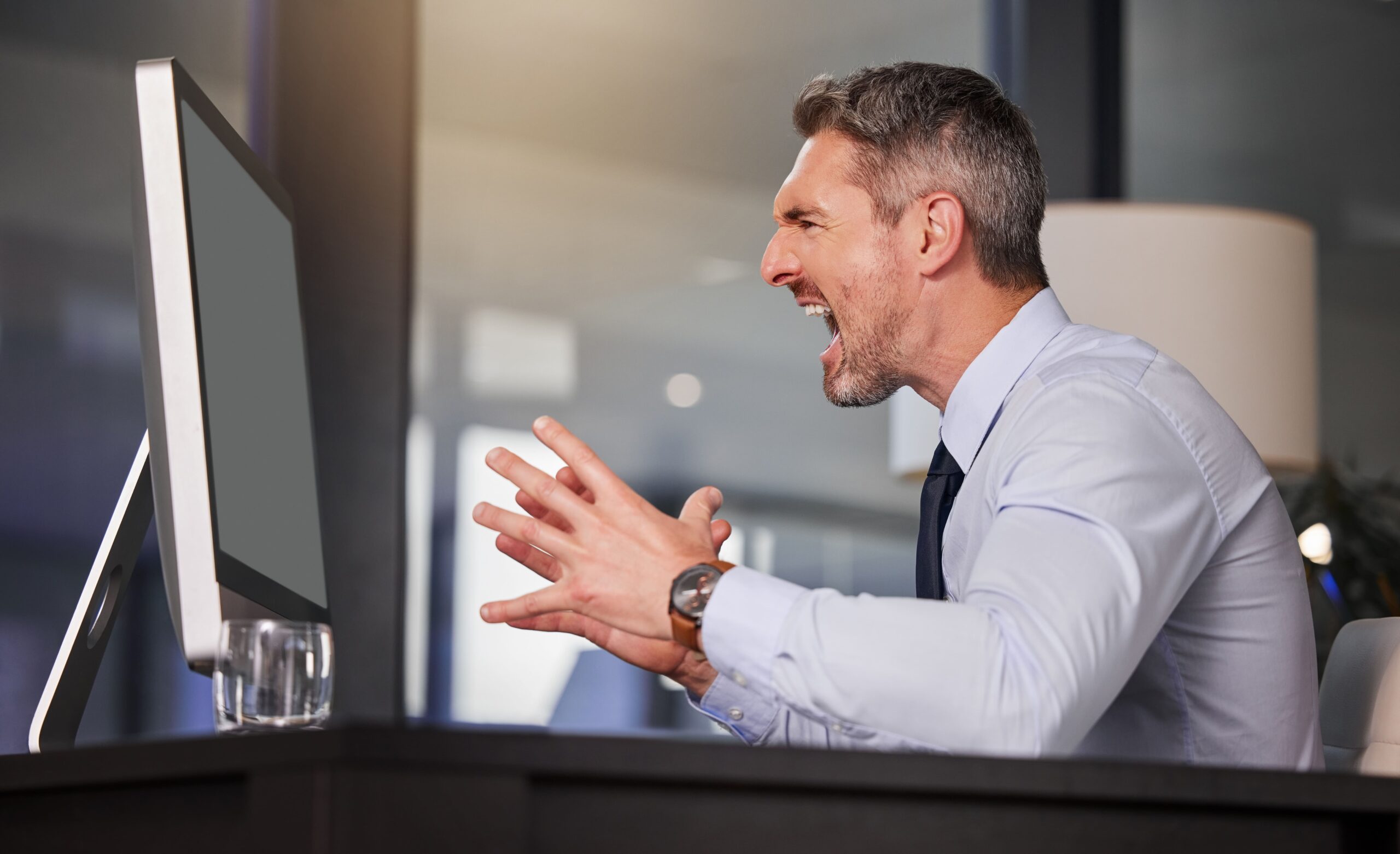 Angry businessman shouting at his computer screen in frustration, representing tech issues, slow performance, or cybersecurity problems at work.