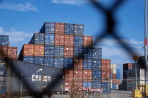 Stacked shipping containers in red, blue, and gray at a distribution yard, viewed through a chain-link fence on a sunny day.