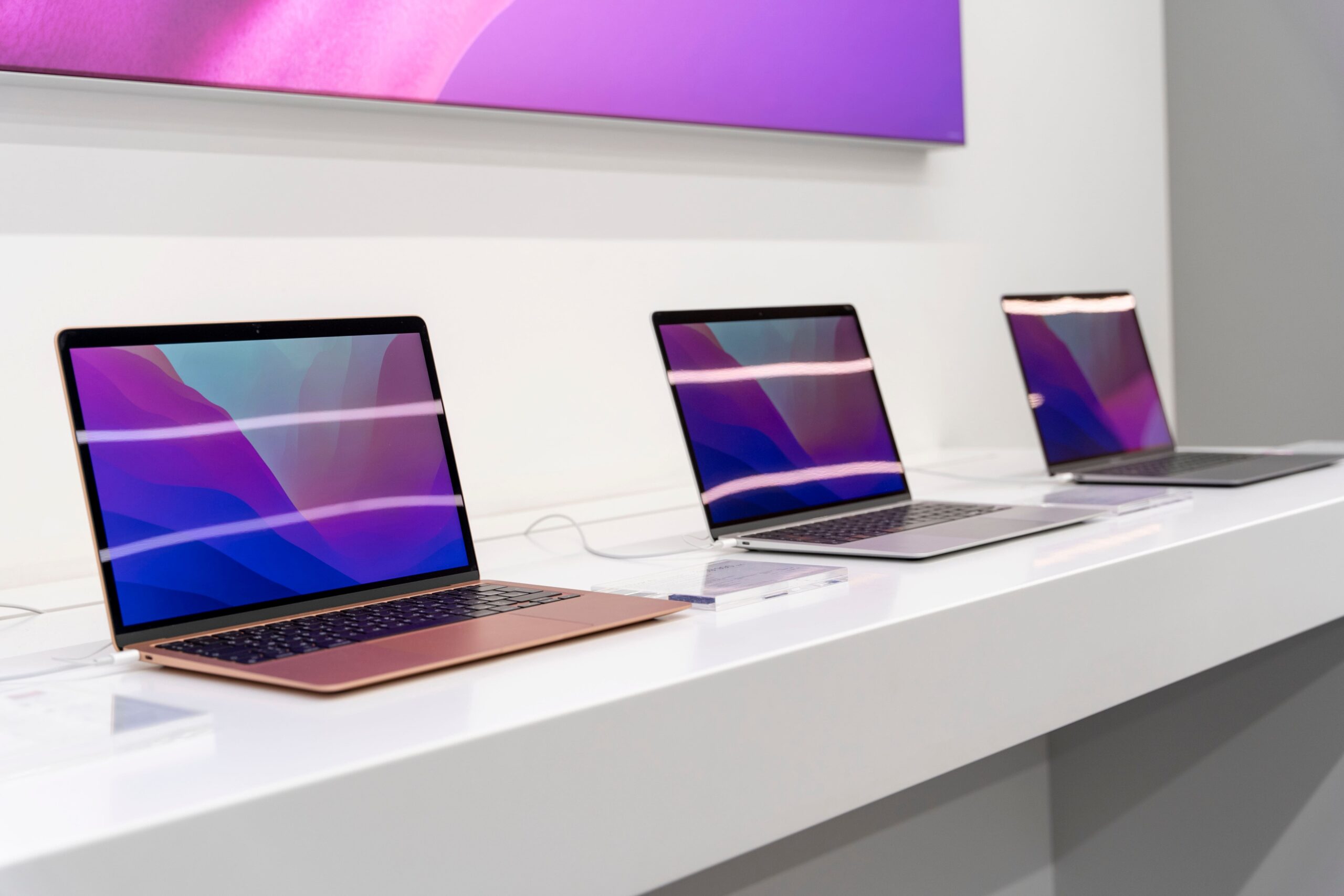 Three modern laptops with purple and pink screens displayed on a white tech store counter beneath.