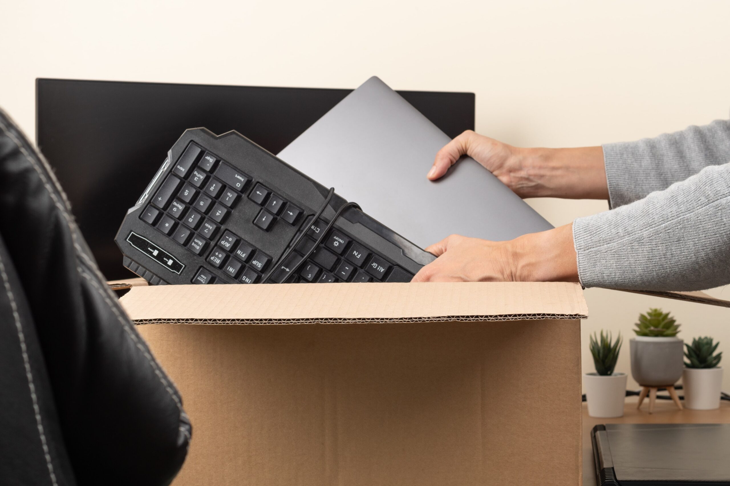 Person packing a computer keyboard and laptop into a cardboard box, suggesting an office upgrade, move, or tech refresh.