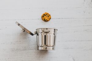 Chocolate chip cookie hovering above a small metal trash can on a white wooden surface, symbolizing internet cookie deletion.