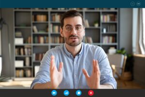Man wearing glasses speaking during a video call, shown on a virtual conference interface with a bookshelf background.