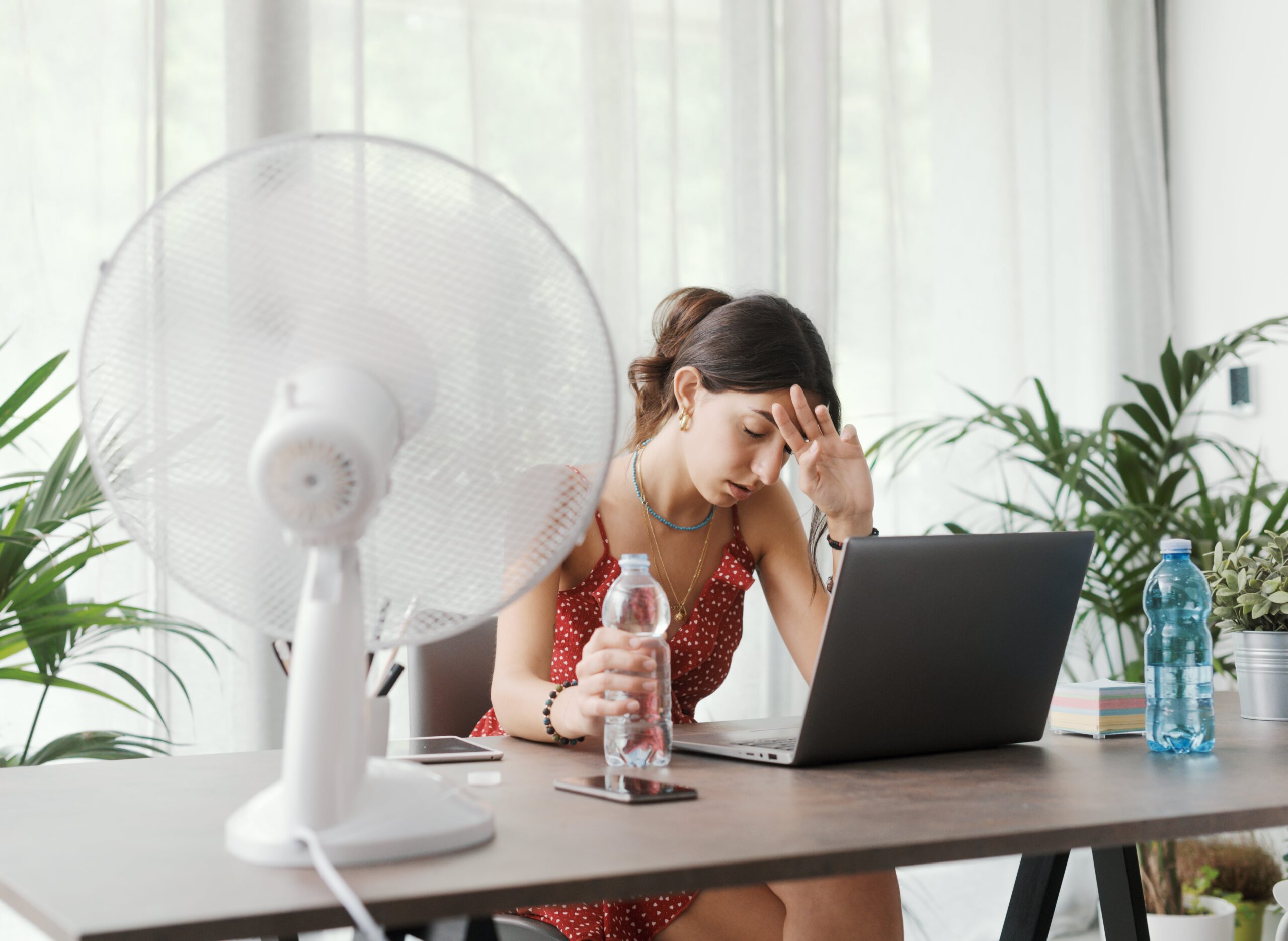 Woman working on a laptop at a desk, visibly hot and tired, holding a water bottle and sitting in front of an electric fan