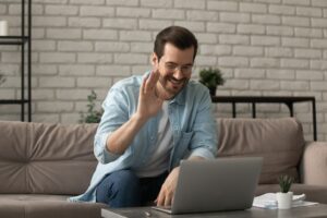 Smiling man sitting on a couch and waving at his laptop during a casual video call at home.
