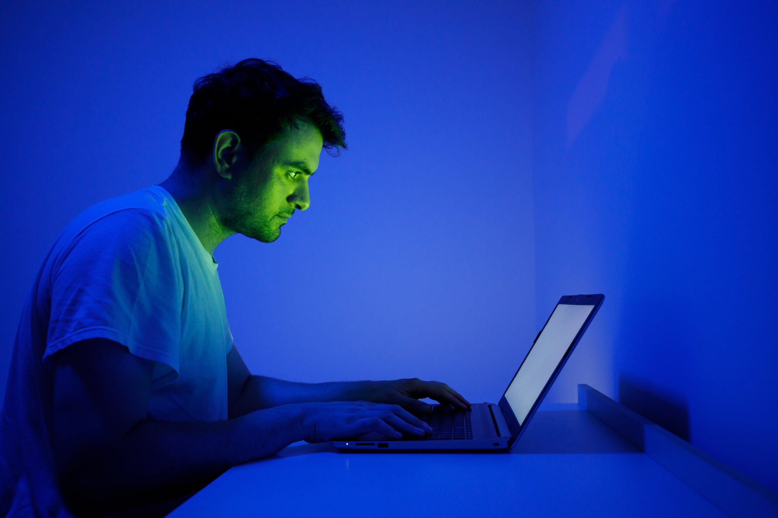 Man using a laptop in a dark room illuminated by a blue and green screen glow, representing late-night computer use or digital eye strain.