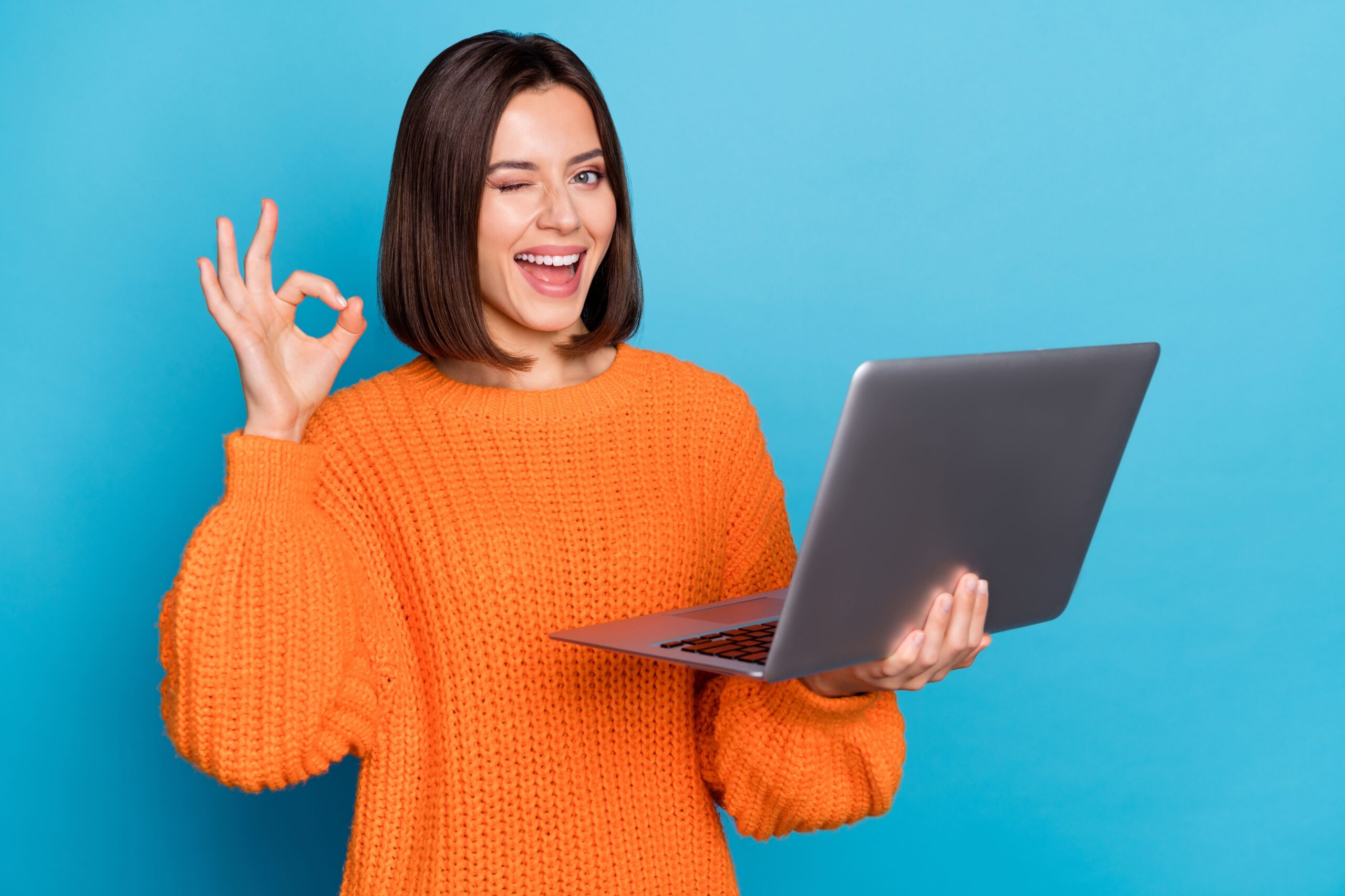 Smiling woman in an orange sweater holding a laptop, winking, and making an OK gesture against a blue background.