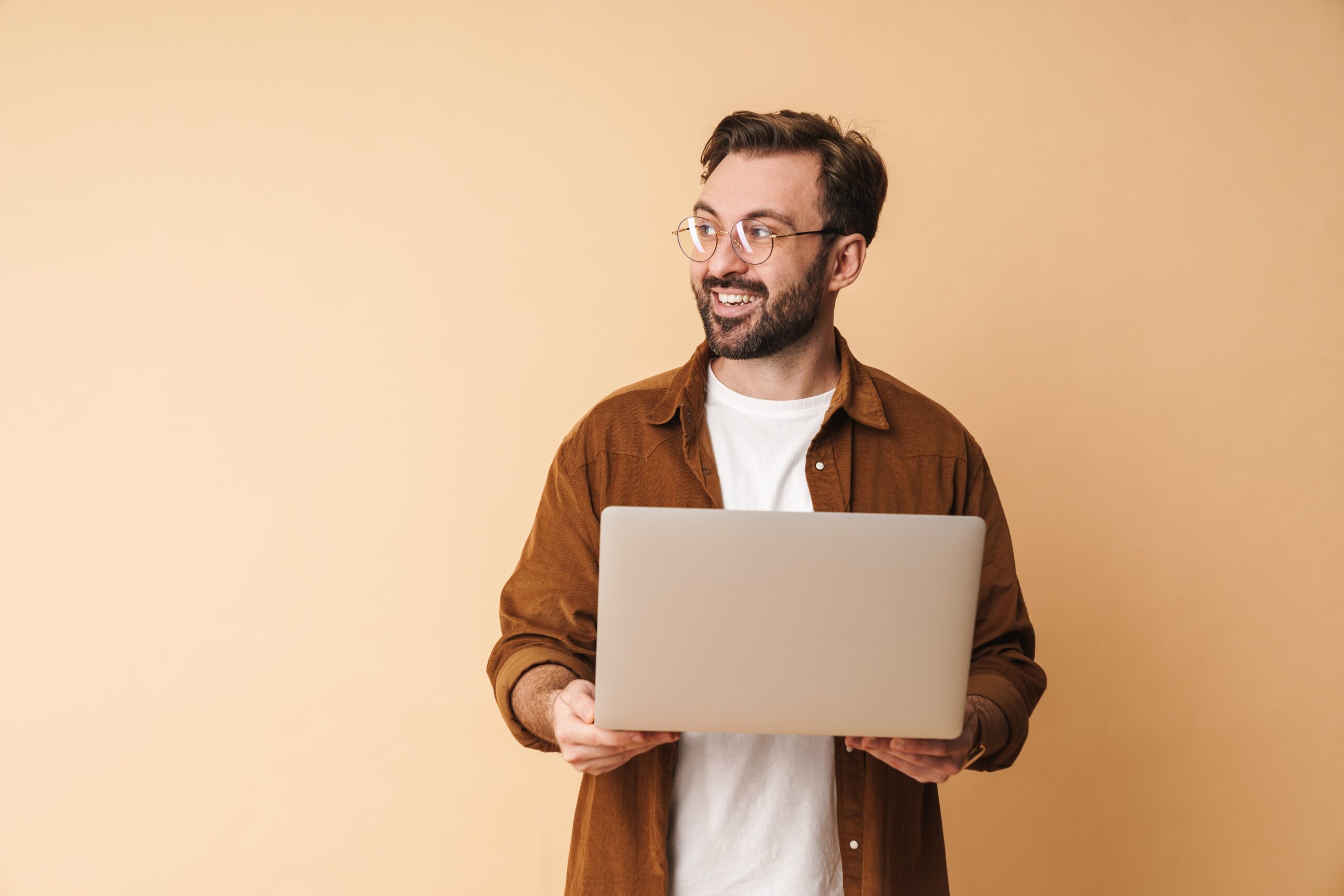 Smiling man wearing glasses and a brown shirt holding an open laptop against a beige background, looking off to the side.