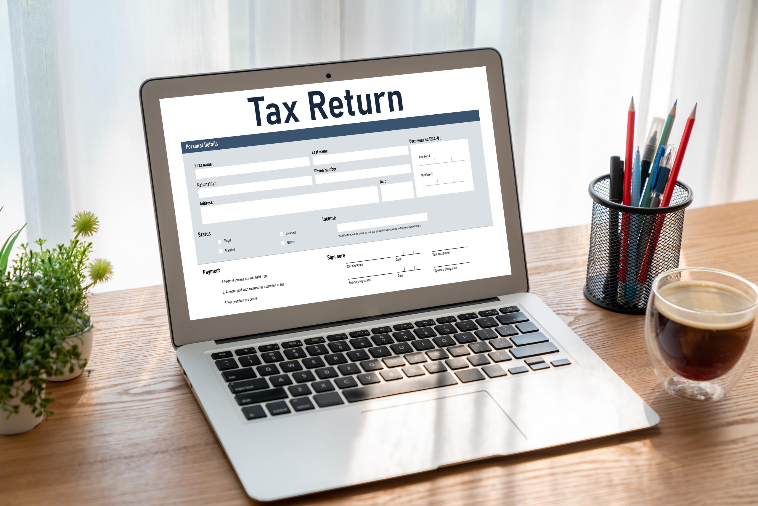 Laptop displaying an online tax return form on a desk, with a coffee cup, pencil holder, and small plant nearby. Natural light filters through sheer curtains in the background.