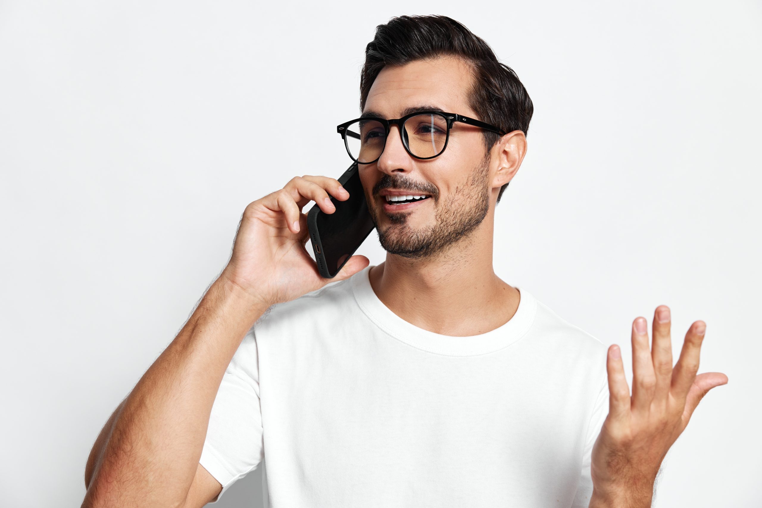 Man wearing glasses talking on a smartphone, smiling and gesturing with his hand against a neutral background.
