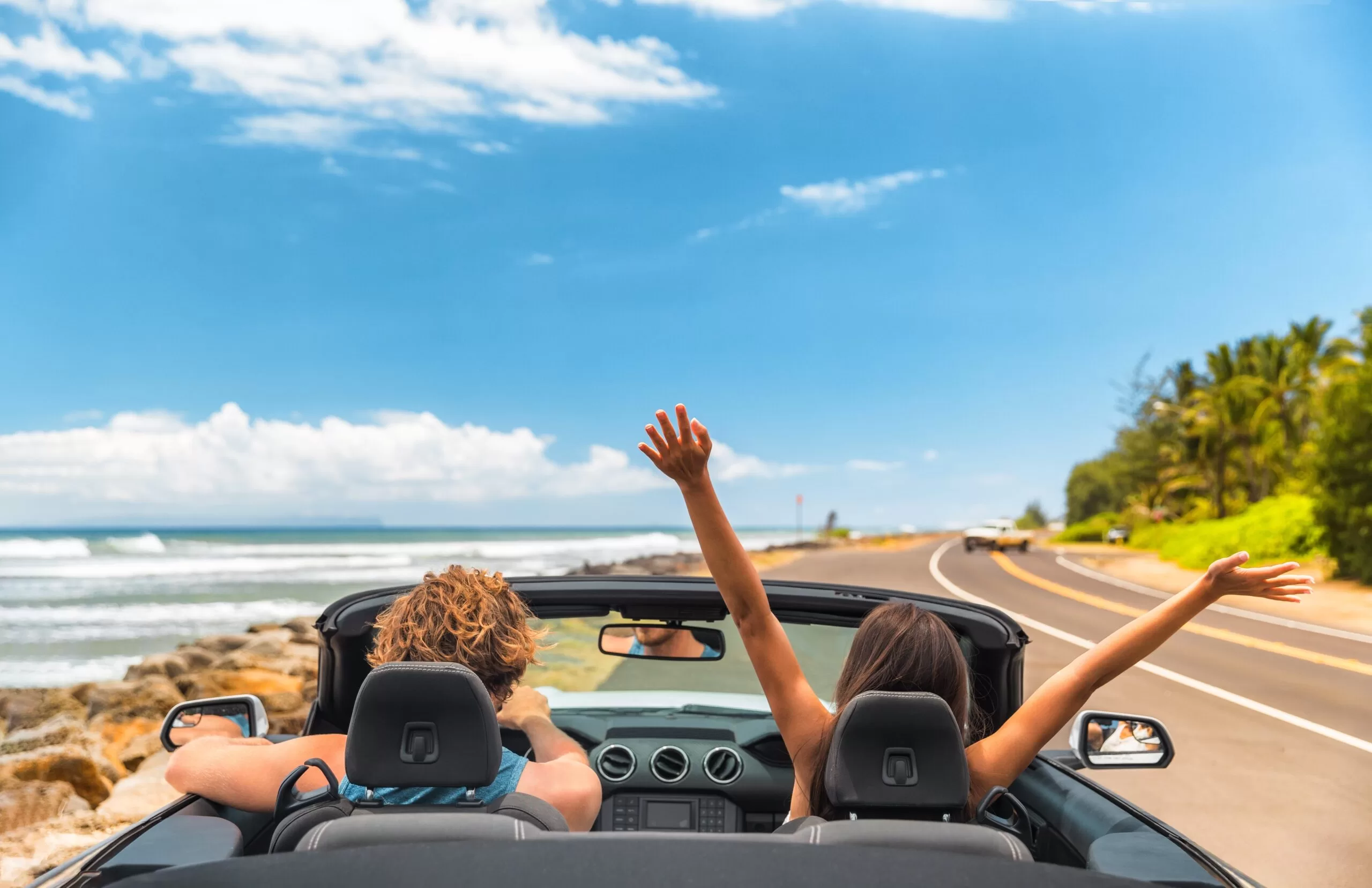 Couple driving convertible car along coastal road with ocean view and hands raised in joy