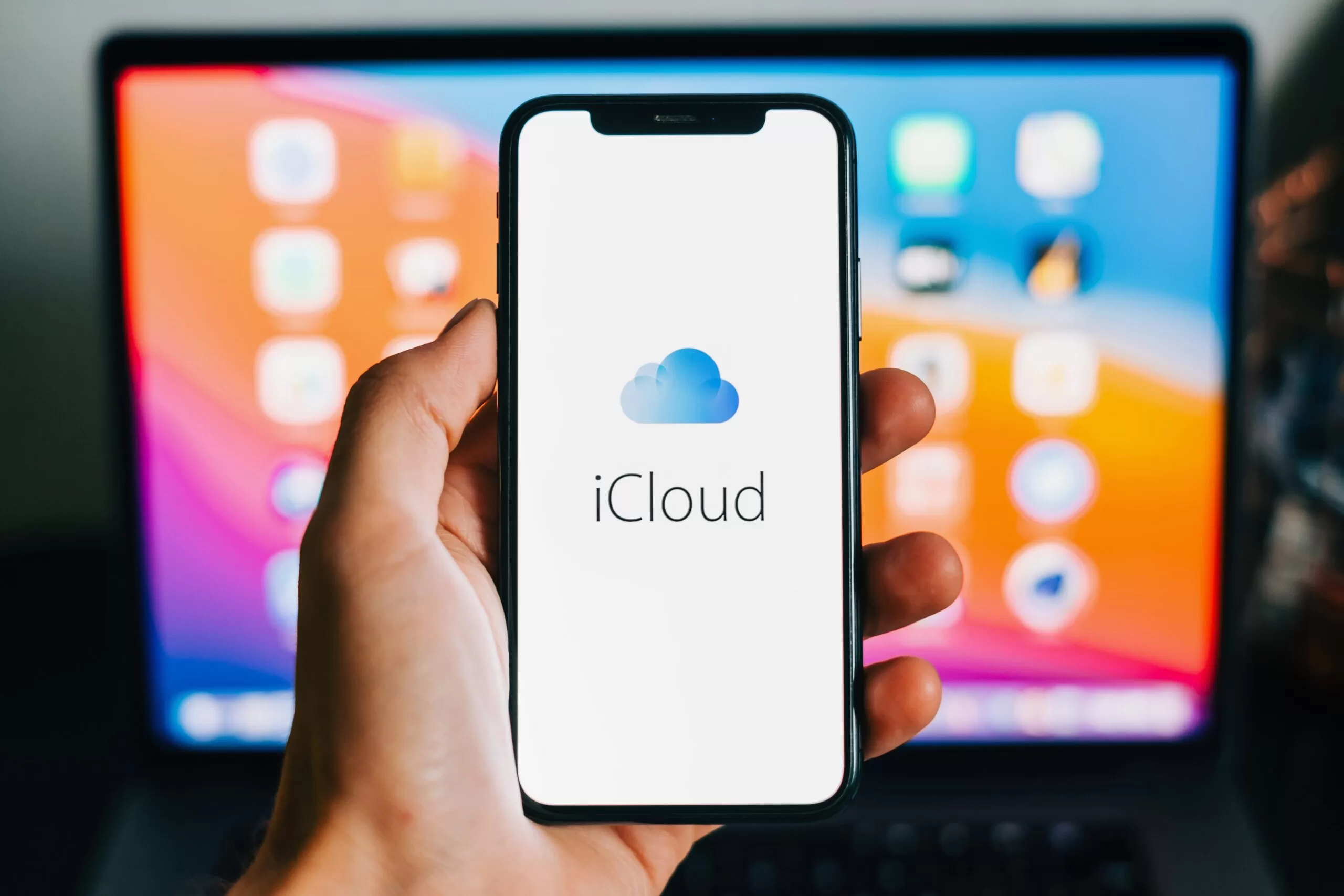 Phone with iCloud logo appearing on screen and Macbook in background.