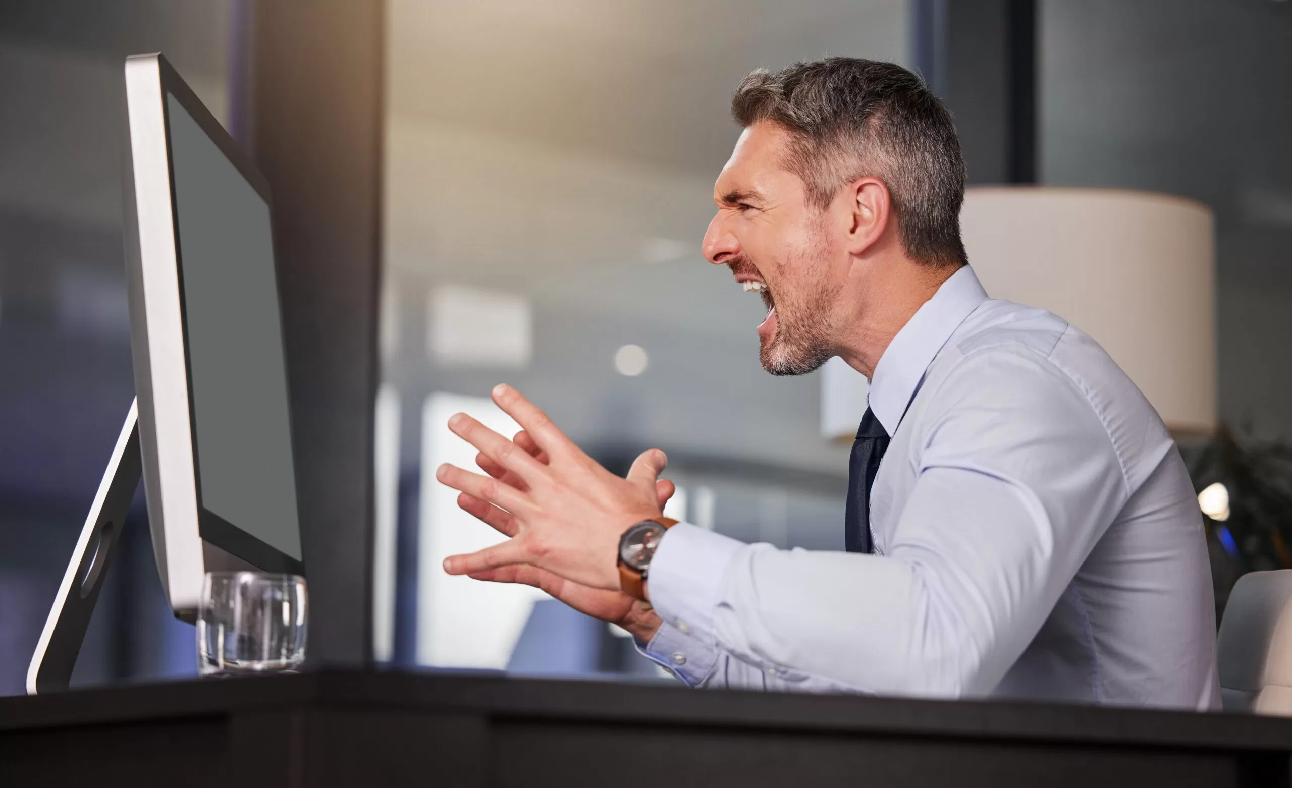 Angry businessman shouting at his computer screen in frustration, representing tech issues, slow performance, or cybersecurity problems at work.