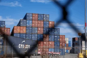 Stacked shipping containers in red, blue, and gray at a distribution yard, viewed through a chain-link fence on a sunny day.