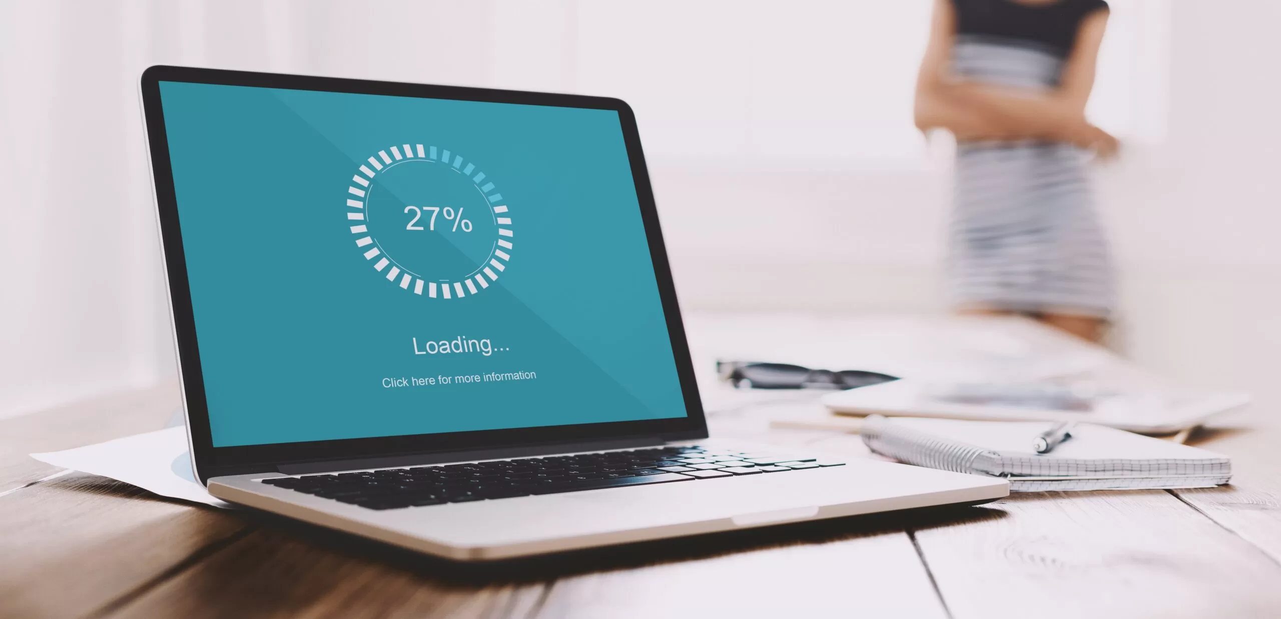 Laptop on a desk displaying a blue loading screen stuck at 27%, suggesting slow performance or system lag, with a blurred person in the background.