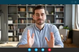 Man wearing glasses speaking during a video call, shown on a virtual conference interface with a bookshelf background.