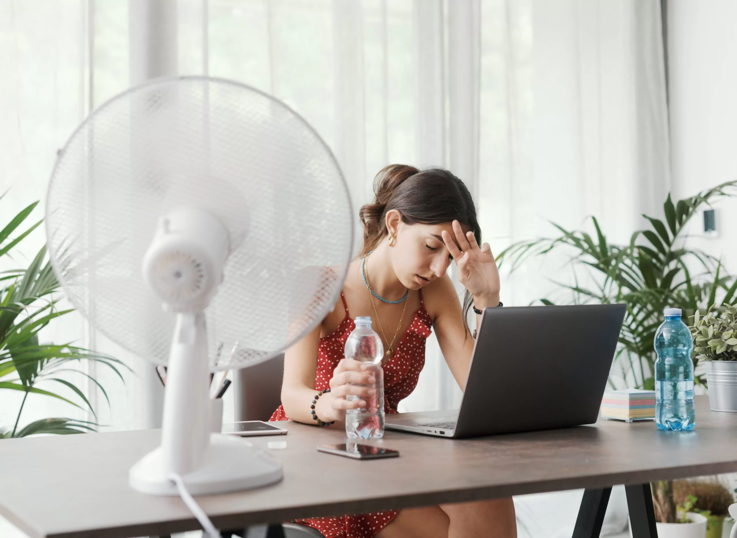 Woman working on a laptop at a desk, visibly hot and tired, holding a water bottle and sitting in front of an electric fan