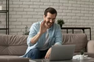 Smiling man sitting on a couch and waving at his laptop during a casual video call at home.