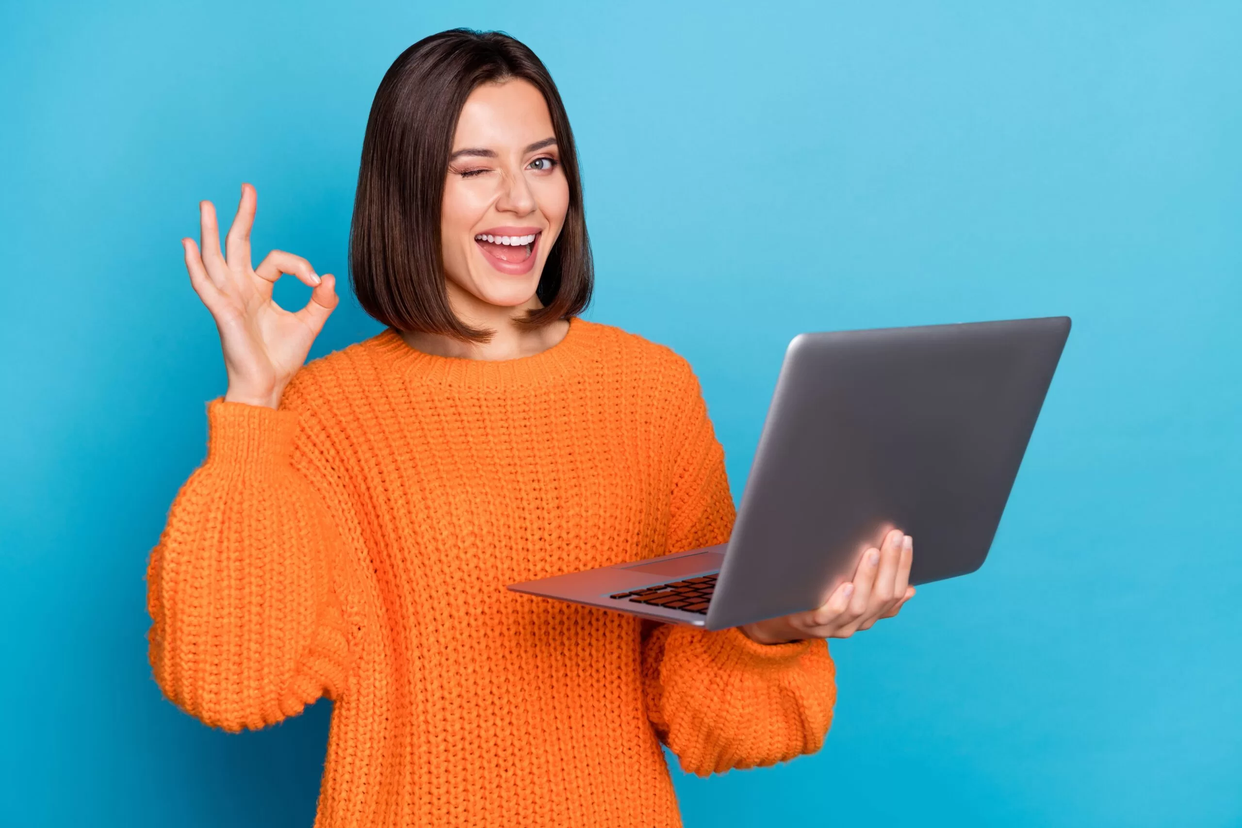 Smiling woman in an orange sweater holding a laptop, winking, and making an OK gesture against a blue background.