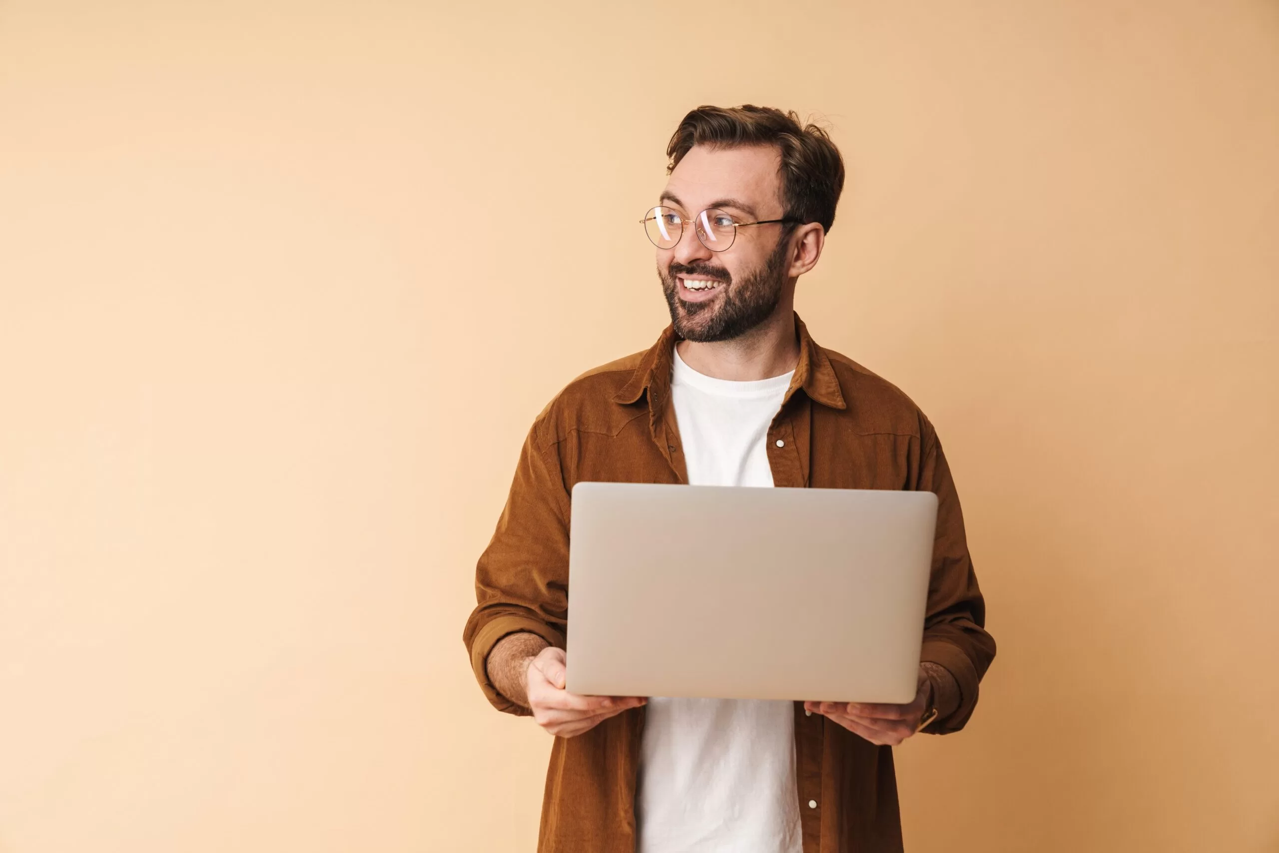 Smiling man wearing glasses and a brown shirt holding an open laptop against a beige background, looking off to the side.