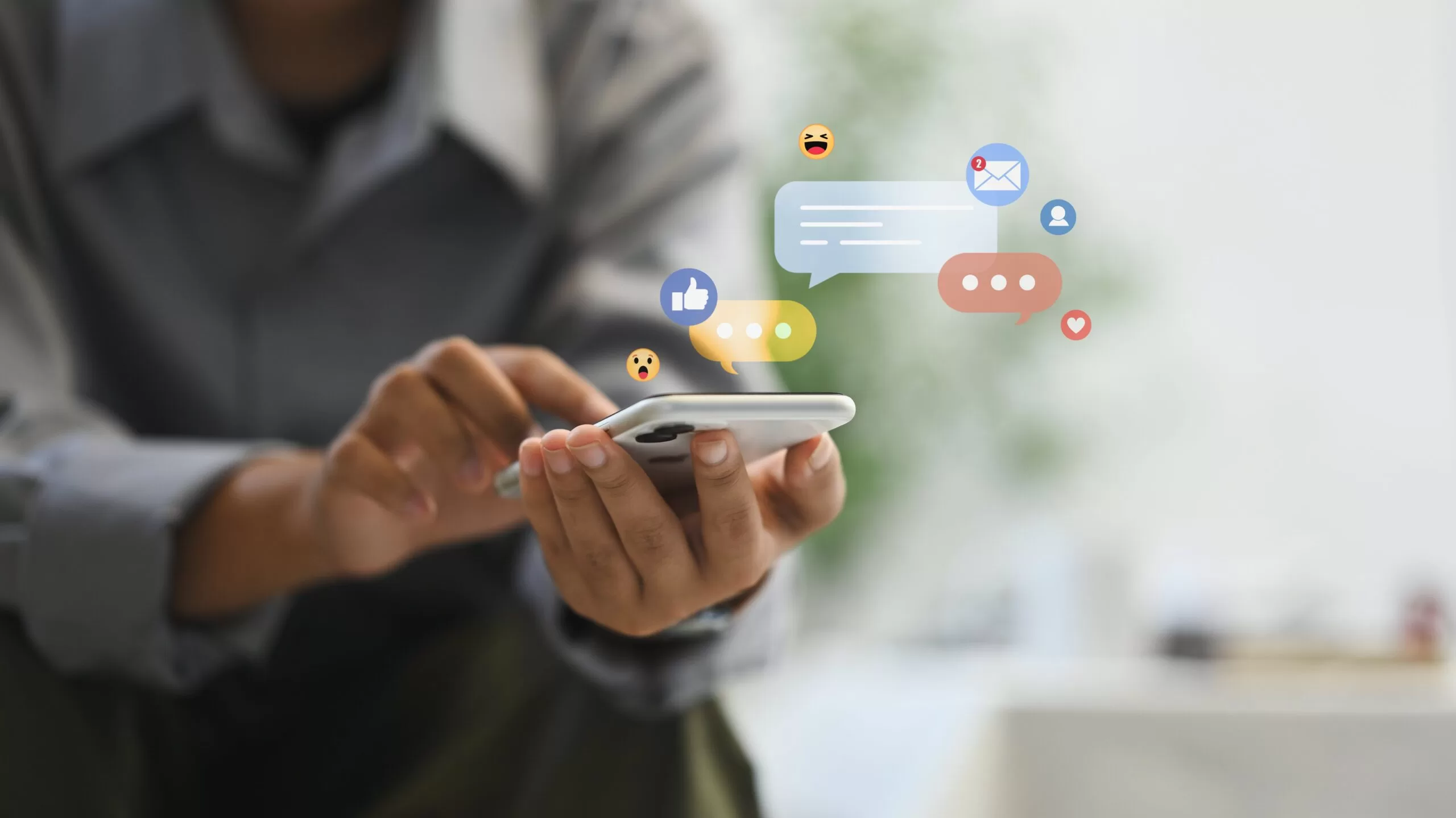 Close-up of a person holding a smartphone with digital icons for likes, messages, emojis, and notifications floating above the screen, symbolizing social media engagement and communication.